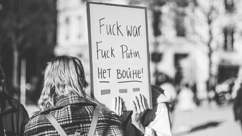 Bild in schwarz weiß gehalten. Frau dreht einem dem Rücken zu, aber Schild von Demonstration ist zu lesen: Fuck war, fuck Putin und dann noch etwas auf Kyrillisch.